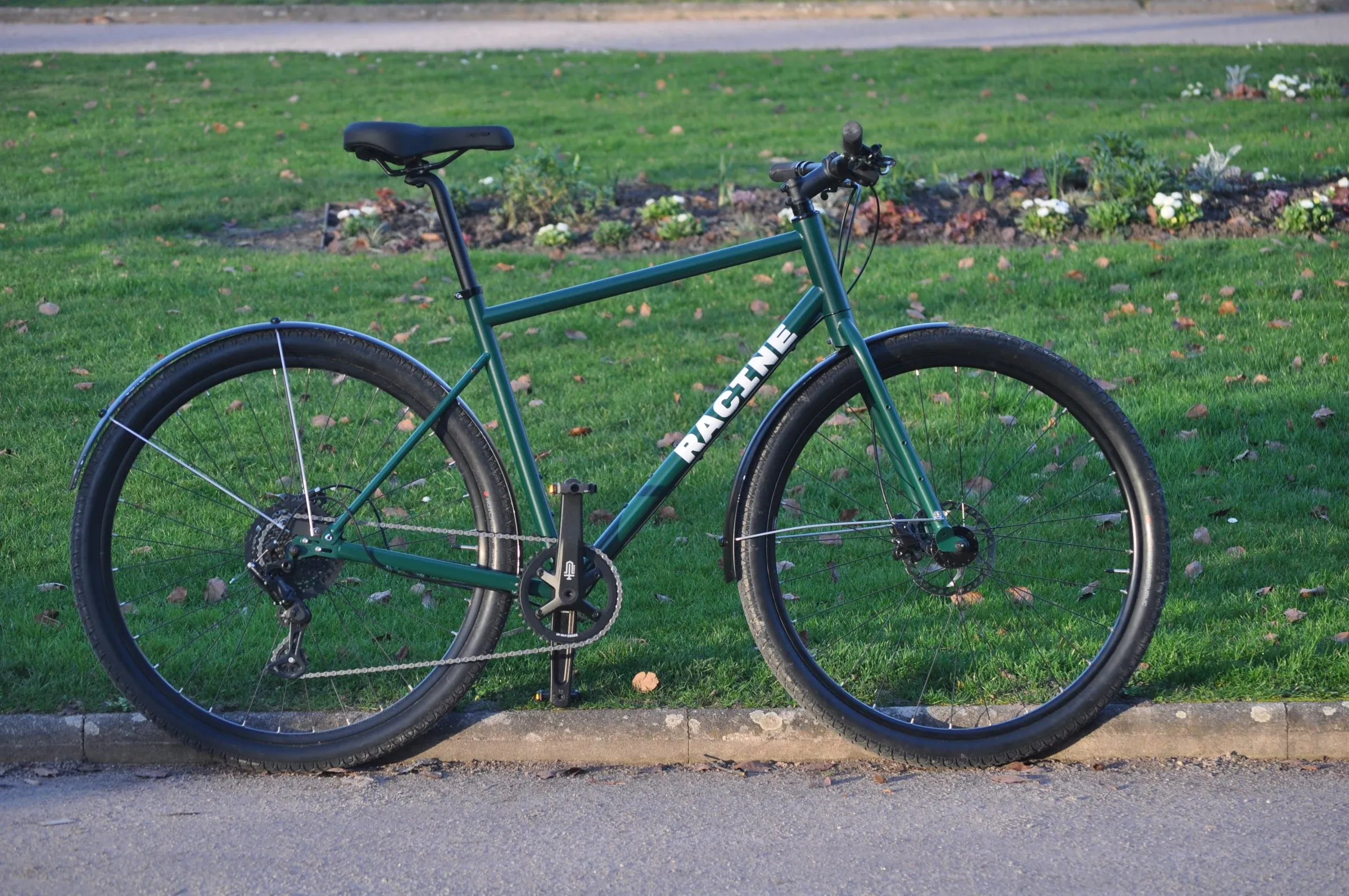 Vélo Racine vert mousse de profil au bord d'une allée de parc, cintre plat et freins à disque